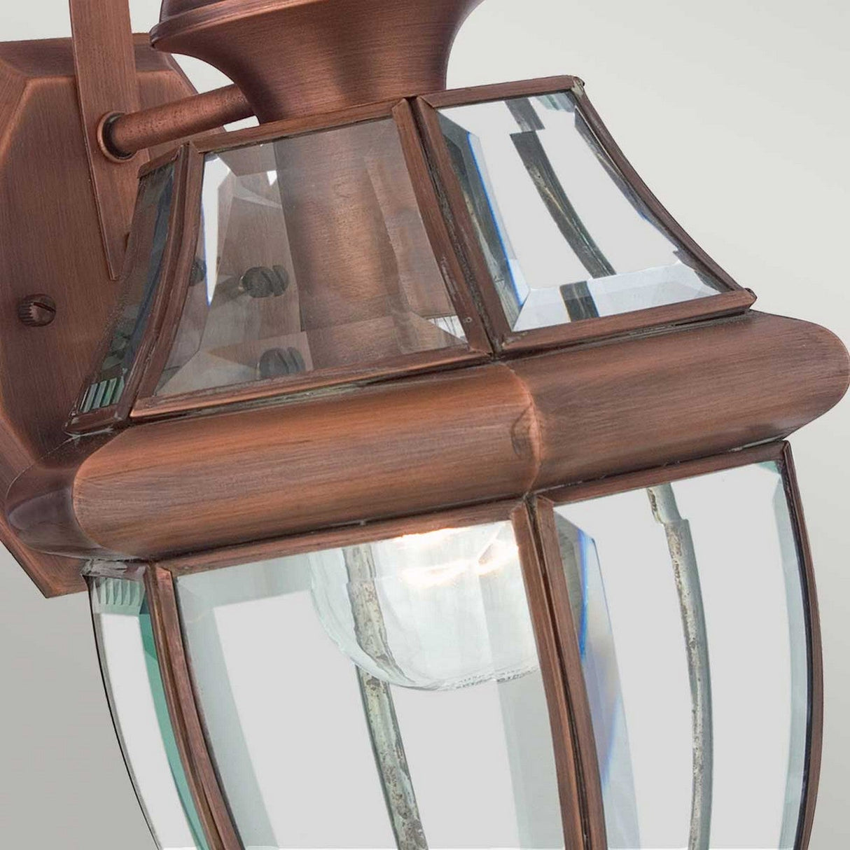 Close-up of the Newbury Outdoor 1 Light Medium Wall Lantern in lacquered aged copper, showcasing its clear beveled glass panels that reveal a visible light bulb inside, beautifully mounted against a light-colored background.