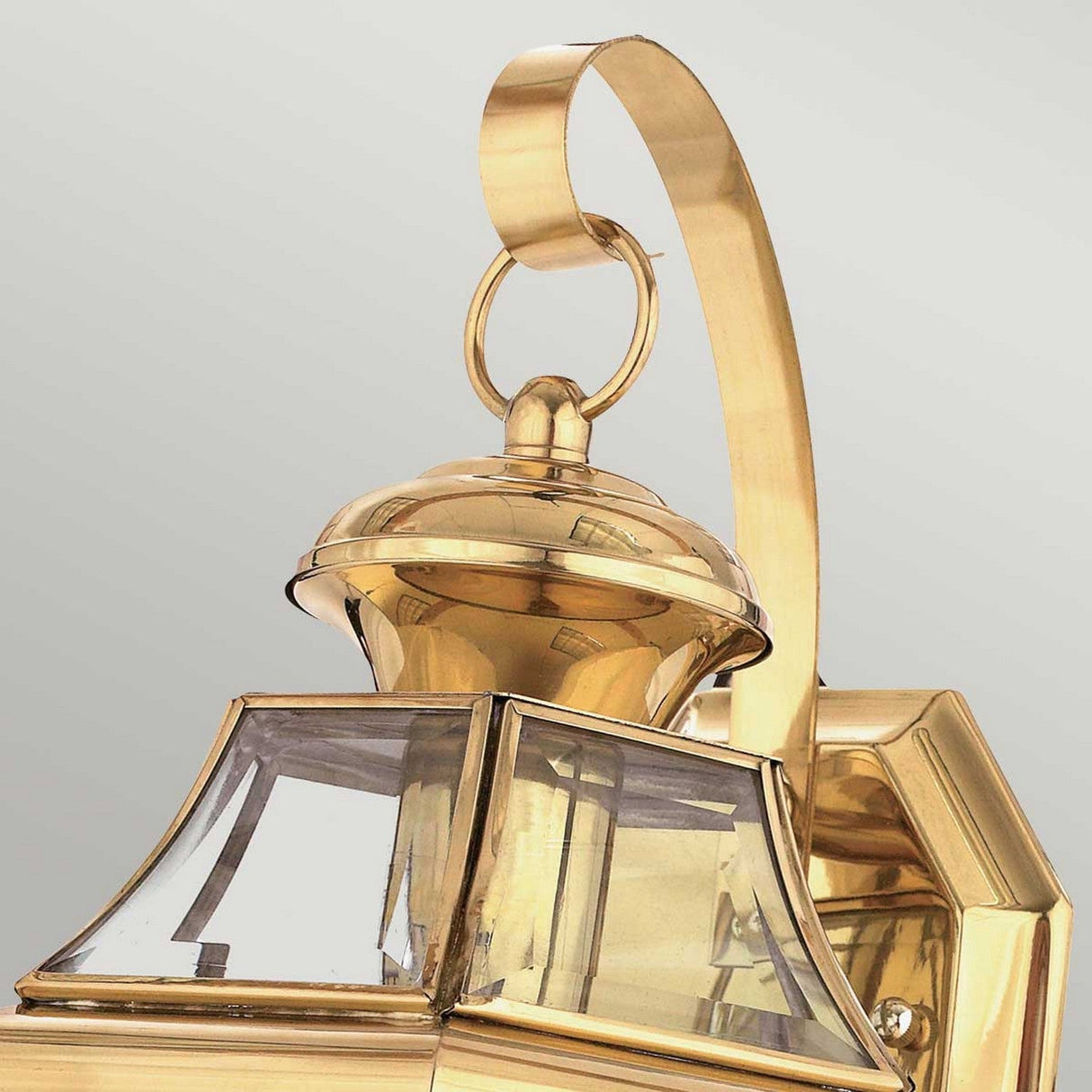 A close-up view of the Newbury Outdoor 1 Light Medium Wall Lantern in lacquered polished brass, showcasing beveled glass panels and a curved handle against a light gray background. This lantern exudes a decorative, vintage style with an elegant, shiny finish.