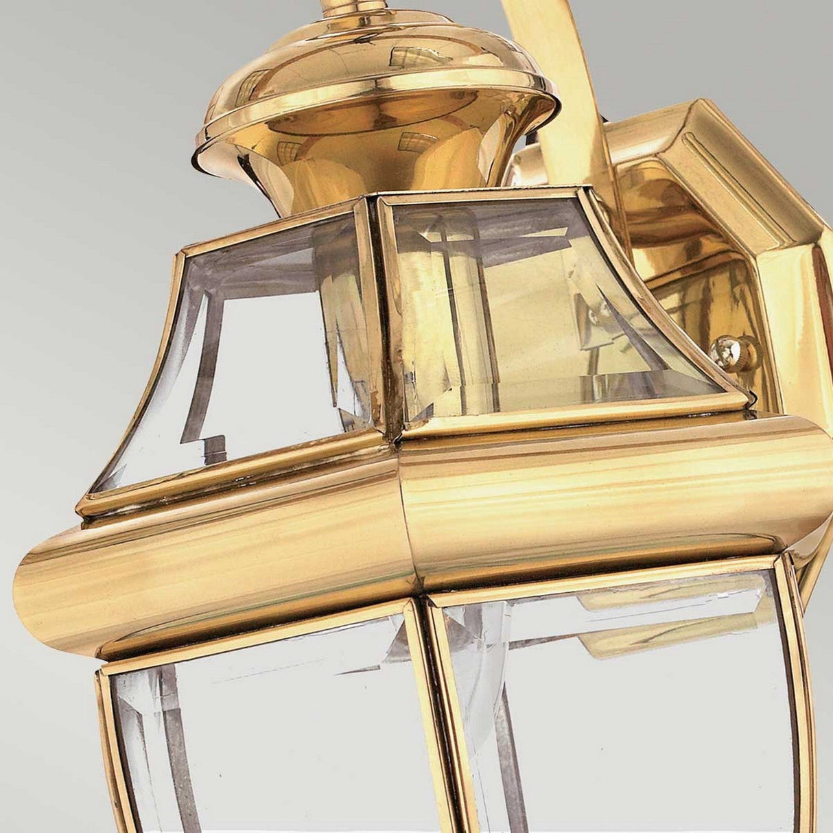 Close-up of the Newbury Outdoor 1 Light Medium Wall Lantern in lacquered polished brass, featuring clear, beveled glass panels, set against a light gray background.