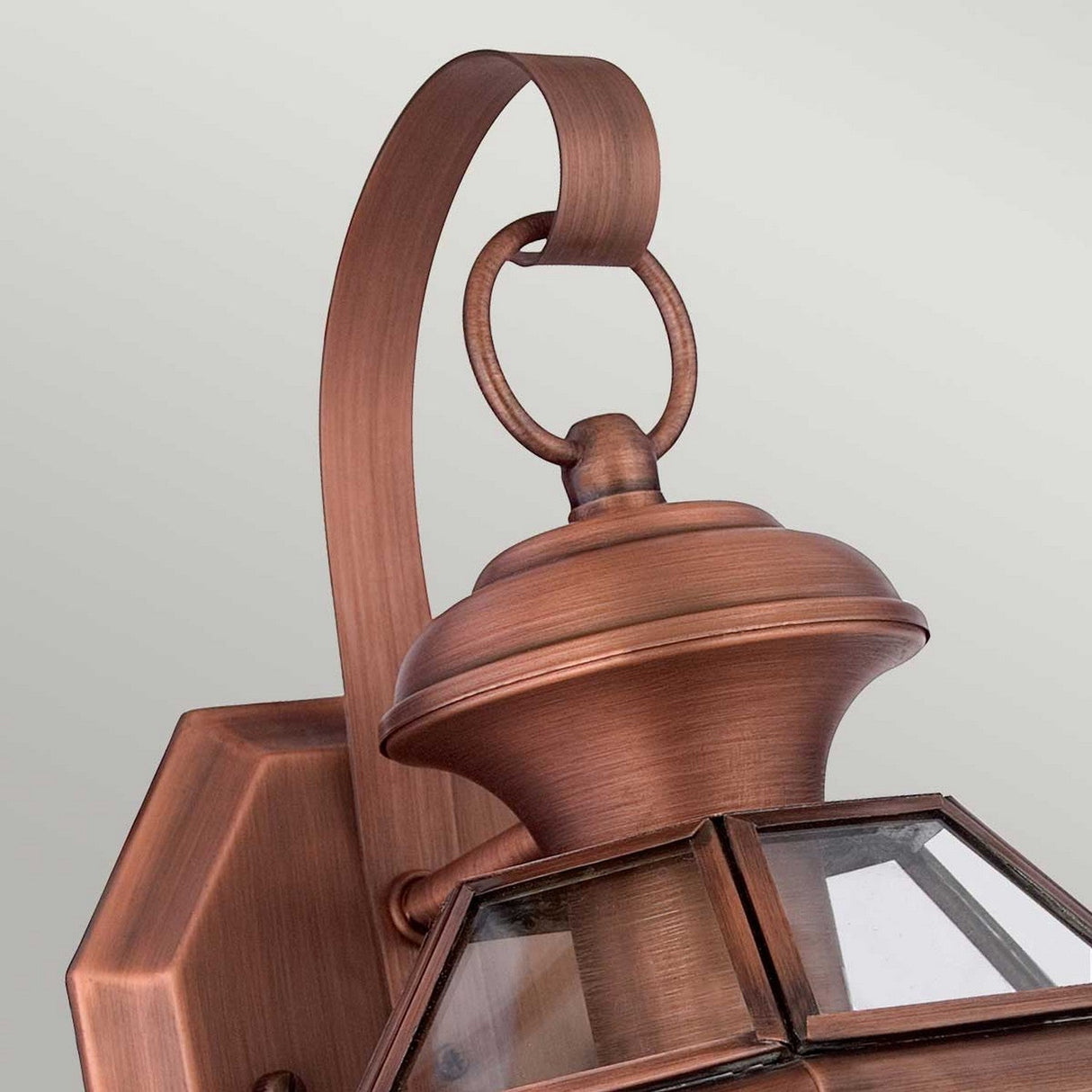 A close-up of the Newbury Outdoor 1 Light Small Wall Lantern reveals its elegant features, including a curved arm and hexagonal clear beveled glass panes. The lacquered aged copper finish beautifully enhances the circular ring detail, adding a sophisticated touch to the fixture.