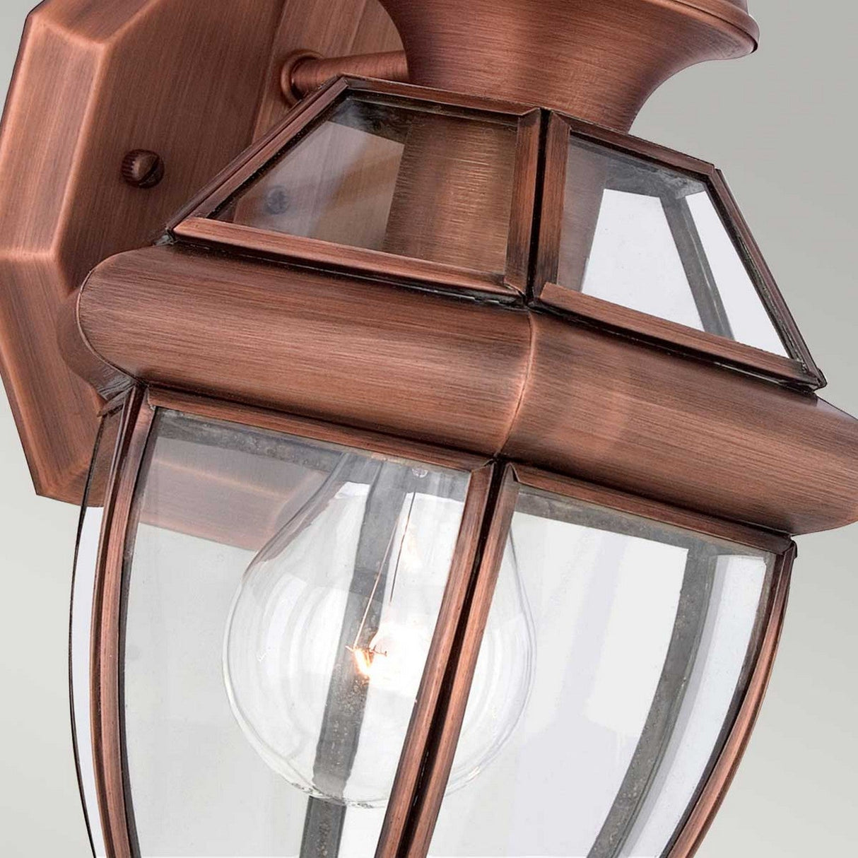 A close-up view of the Newbury Outdoor 1 Light Small Wall Lantern reveals its elegant design with clear beveled glass panels, showcasing the visible light bulb inside and highlighting the lacquered aged copper finish.