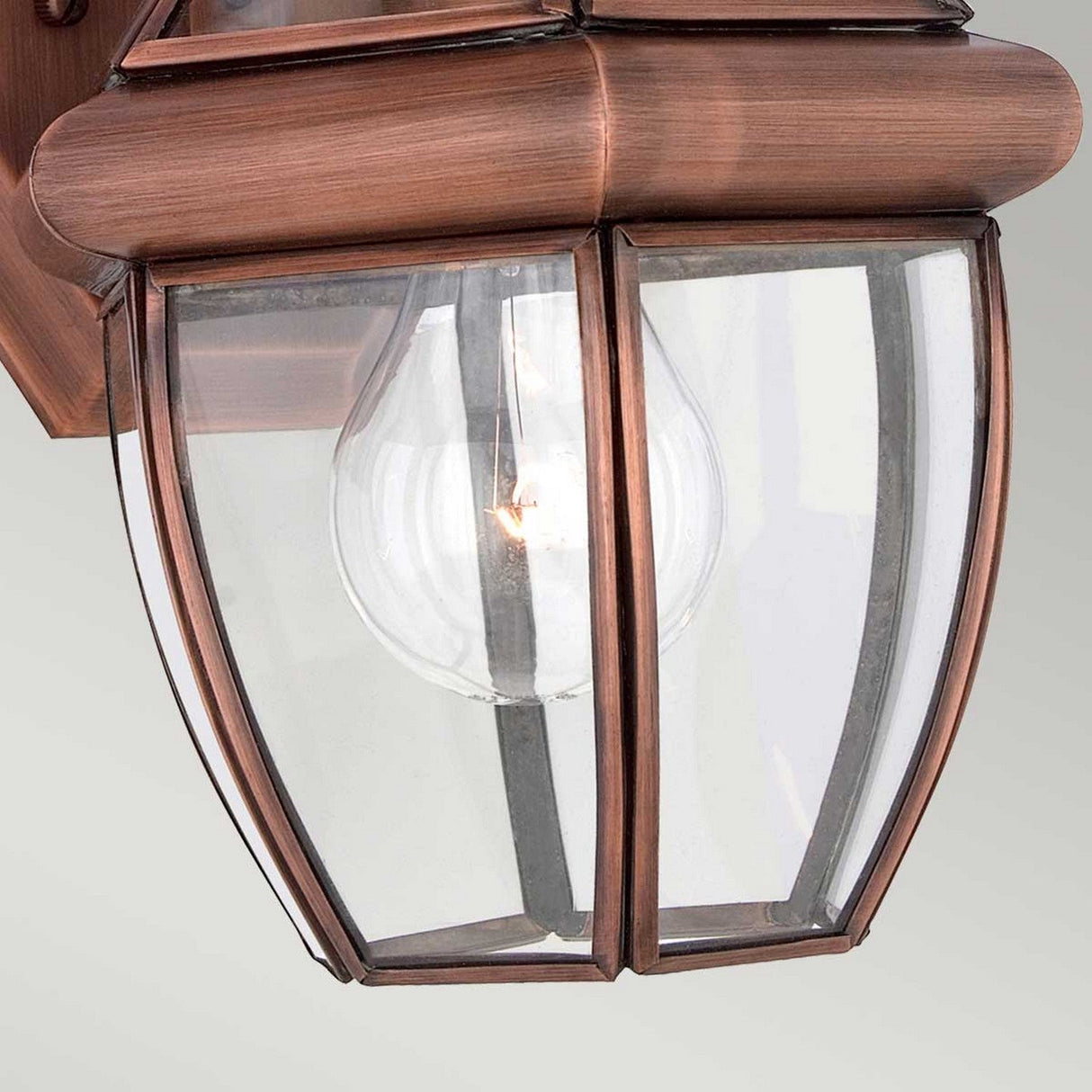 Close-up of the Newbury Outdoor 1 Light Small Wall Lantern, featuring clear beveled glass and a glowing incandescent bulb inside. The fixture is finished in lacquered aged copper, offering a classic and elegant design.