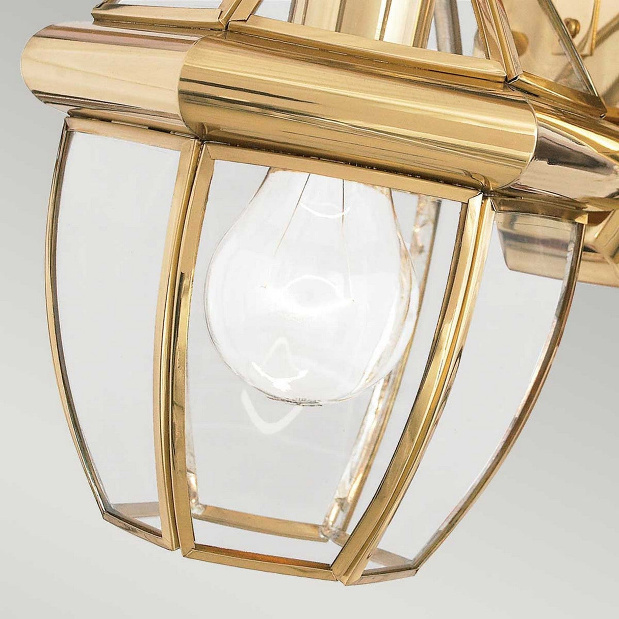 A detailed view of a Newbury Outdoor 1 Light Small Wall Lantern in lacquered polished brass highlights a visible light bulb encased in beveled glass panels. The modern and elegant design features clean lines against a neutral background.
