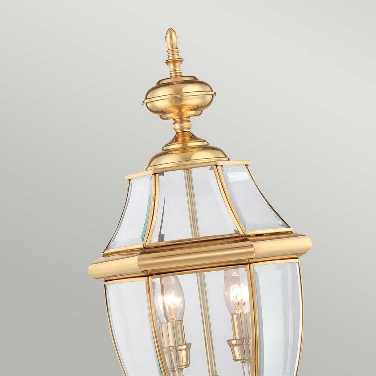 Close-up of the Newbury 2 Light Pedestal in lacquered polished brass, showcasing an elegant classic lantern design with two visible bulbs. The brass frame features decorative elements, and the clear glass panels allow light to shine through—making it a perfect addition to your outdoor lighting setup.