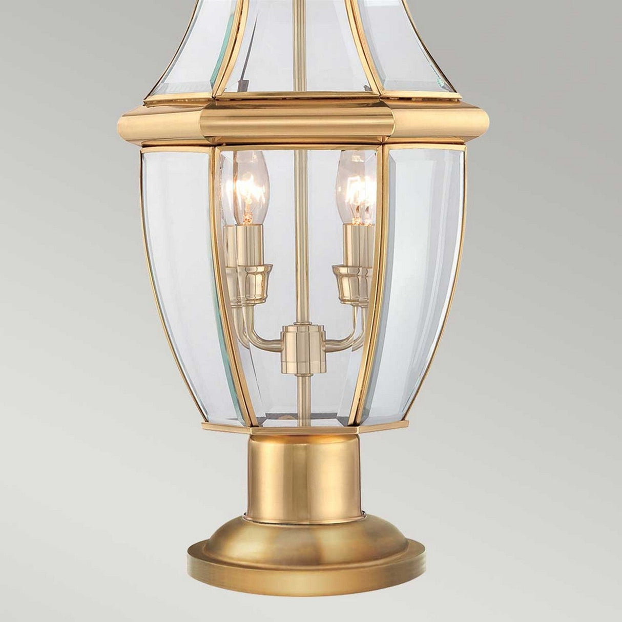 The Newbury 2 Light Pedestal, crafted with clear glass panels and boasting two elegant candle-style bulbs, features a classic design with a warm, lacquered polished brass finish reminiscent of traditional street lamps. This sophisticated piece of outdoor lighting is perfect for adding elegance to any plain background.