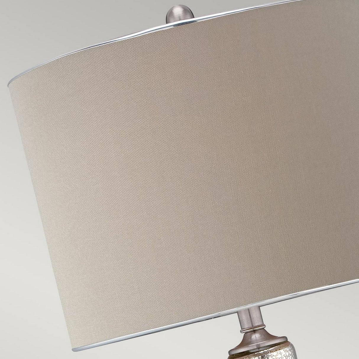A close-up of the Nicolls 1 Light Table Lamp - Mercury Glass showcases a beige lampshade with a sleek metallic base, epitomizing elegance. The image accentuates the fabric texture and top part of the lamp, set against a neutral background that enhances its ambient lighting.