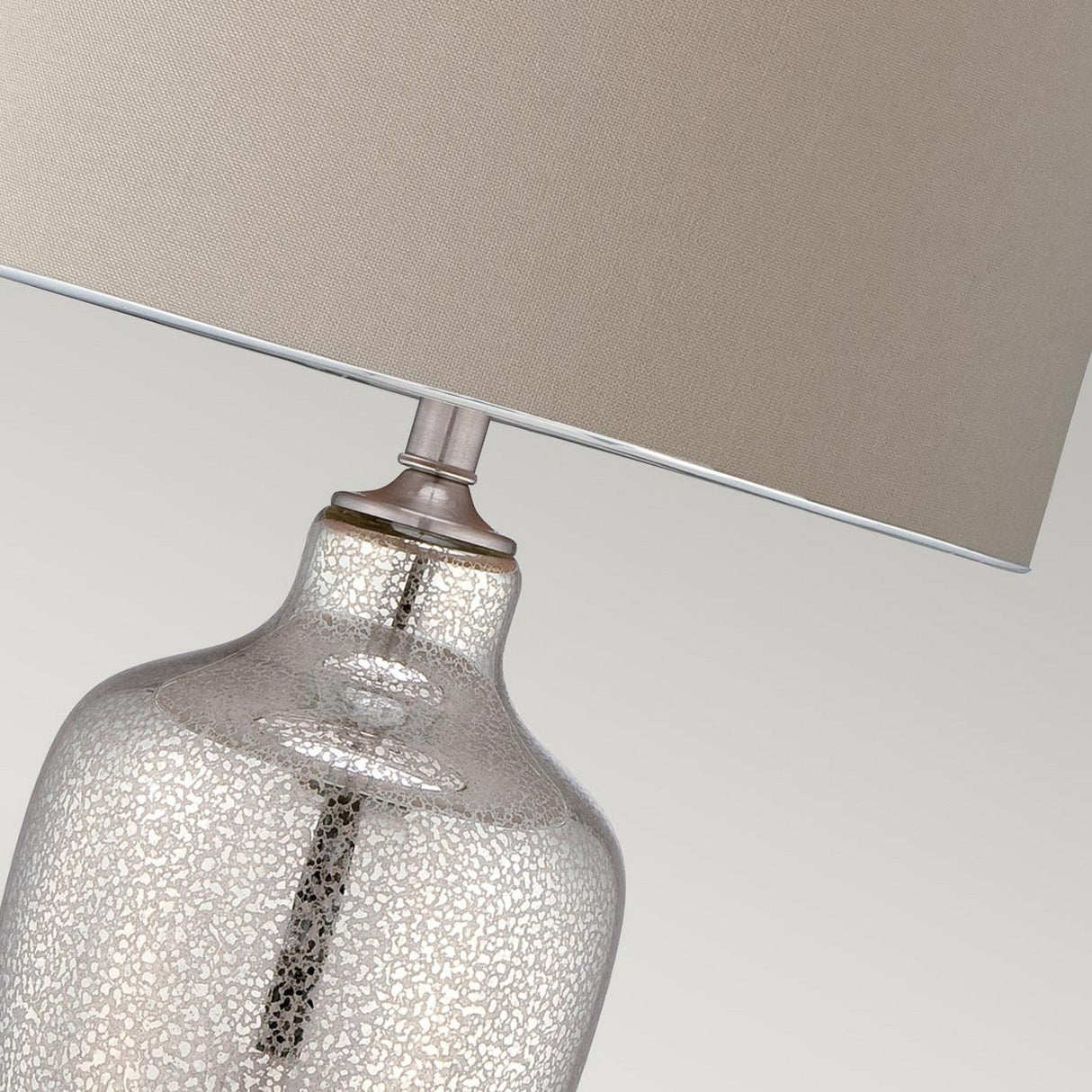 A close-up of the Nicolls 1 Light Table Lamp showcases its elegant design with a textured glass base and beige lampshade. The base features a stunning cracked Mercury glass pattern that beautifully reflects ambient lighting, set against a neutral gray background to enhance its sophisticated appeal.