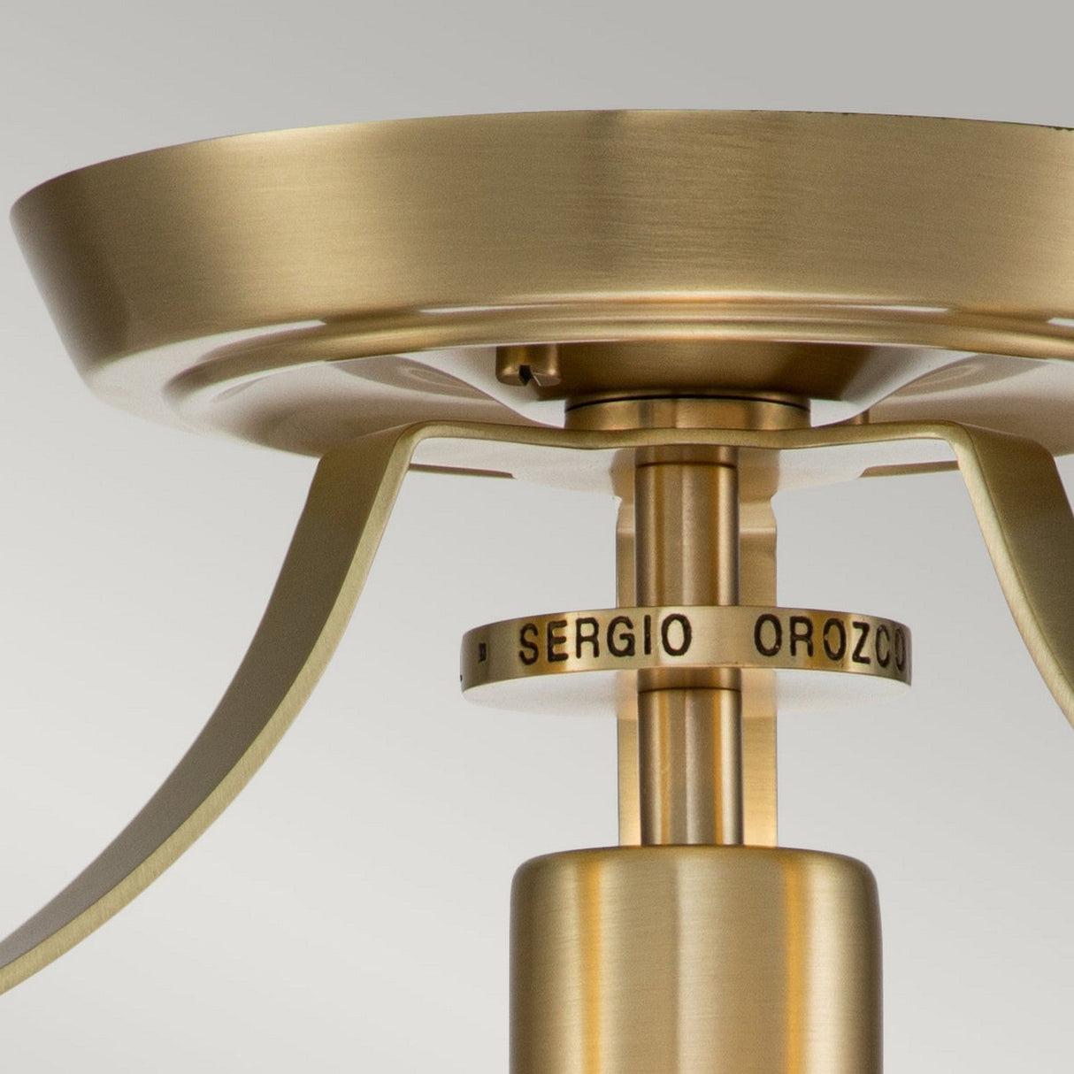 A close-up of the Penn Station 1 Light Semi-Flush in brushed brass showcases a circular base and elegant curved arms extending from the center, while a metal ring features an industrial design inscribed with "SERGIO OROZCO.