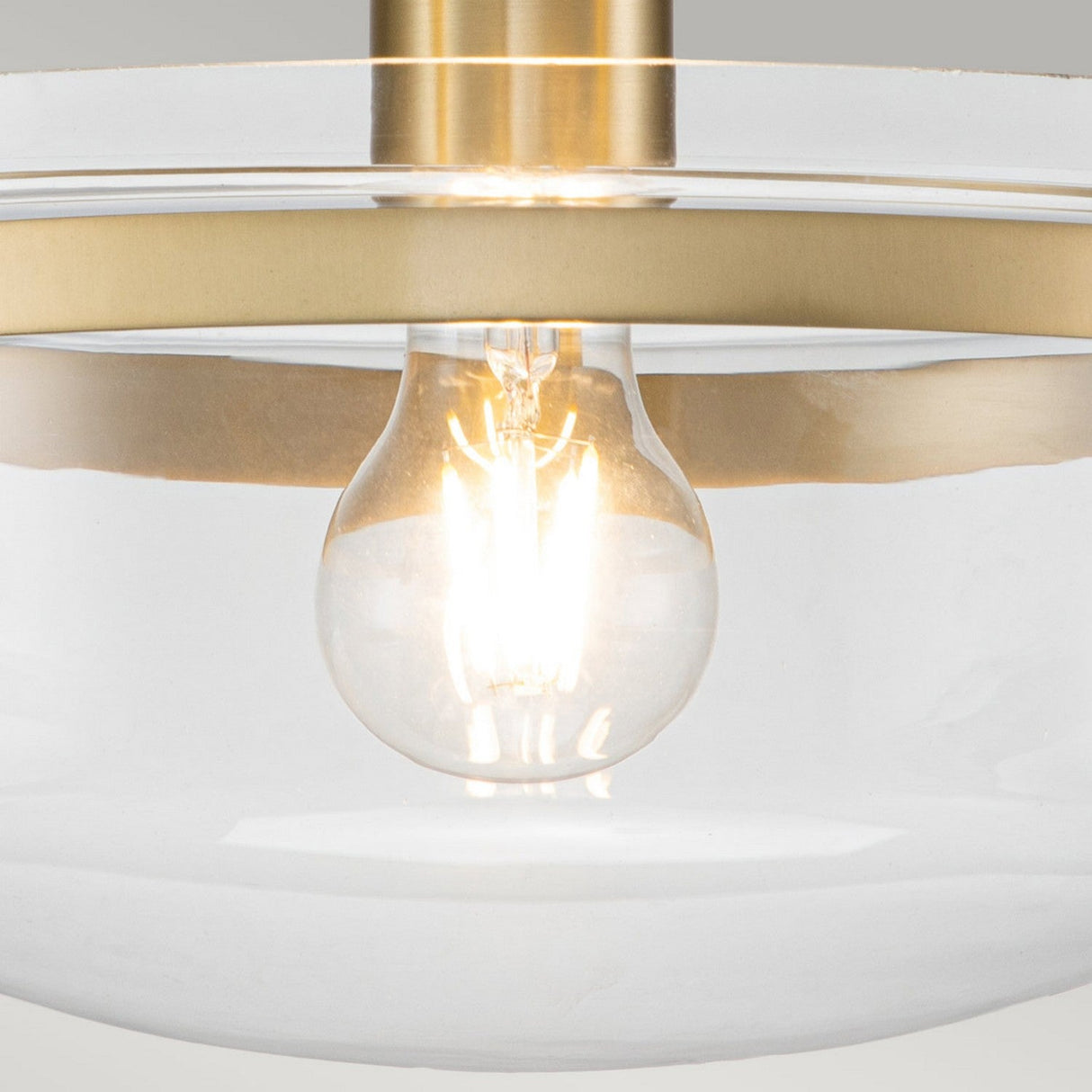 A close-up of the Penn Station 1 Light Semi-Flush in brushed brass highlights its modern design, featuring a clear glass shade through which a glowing bulb is visible. The fixture embodies industrial design elements, giving it a sleek and contemporary appearance.
