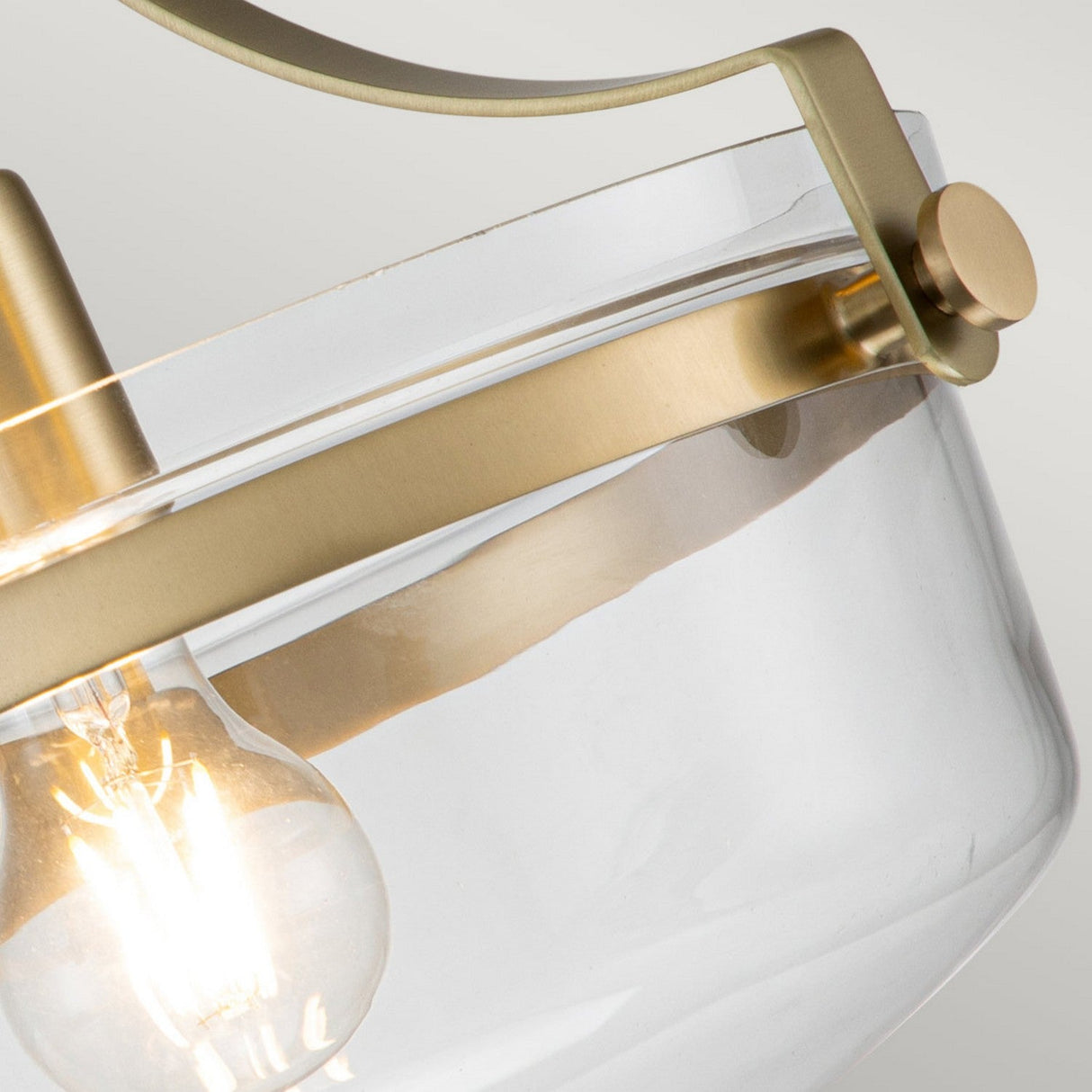 Close-up of the Penn Station 1 Light Semi-Flush in Brushed Brass, showcasing a clear glass shade with a brushed brass metal frame. Illuminated from within to cast a warm glow, its sleek and minimalist design hints at an industrial aesthetic, making it a stylish addition to any contemporary space.