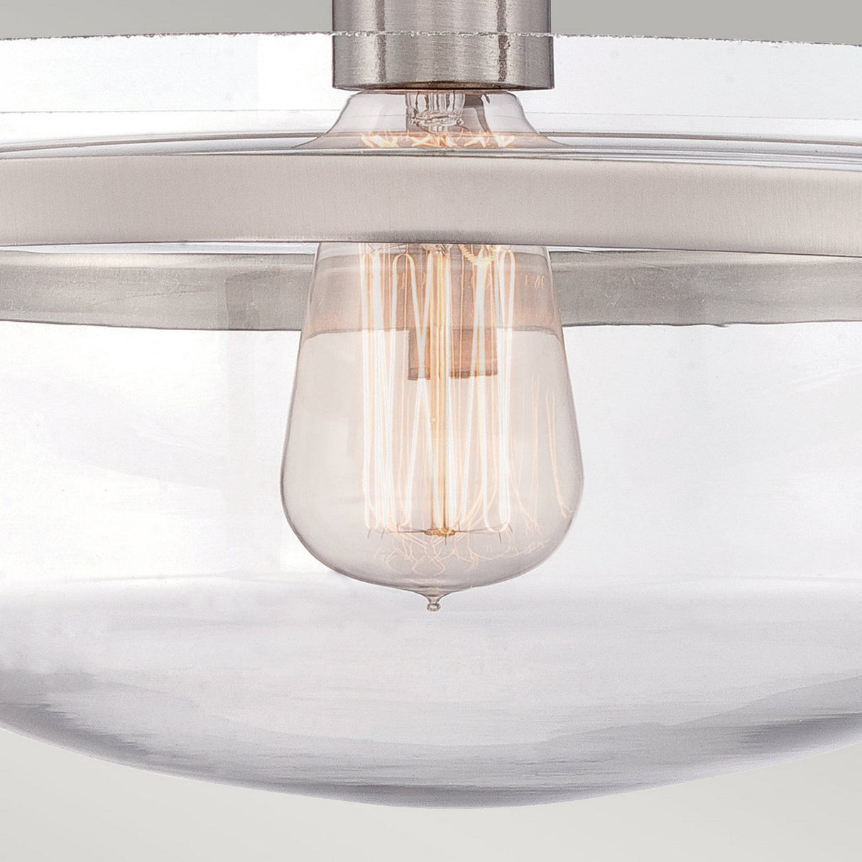 A close-up image showcases the Penn Station 1 Light Semi-Flush in Brushed Nickel, featuring a vintage-style Edison bulb in a clear glass light fixture that exudes industrial charm. The visible glowing filament is beautifully complemented by brushed nickel fixtures securing the glass. The background is a neutral color.