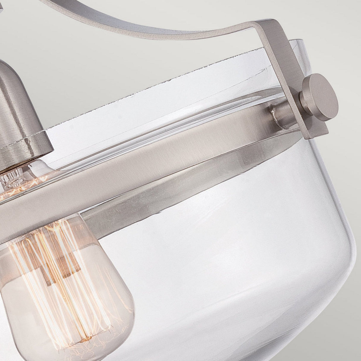 A detailed view of the Penn Station 1 Light Semi-Flush in Brushed Nickel showcases its industrial charm, highlighted by a clear glass shade and visible filament bulb. The design features a sleek metallic structure with minimalist elements characteristic of the Penn Station aesthetic.