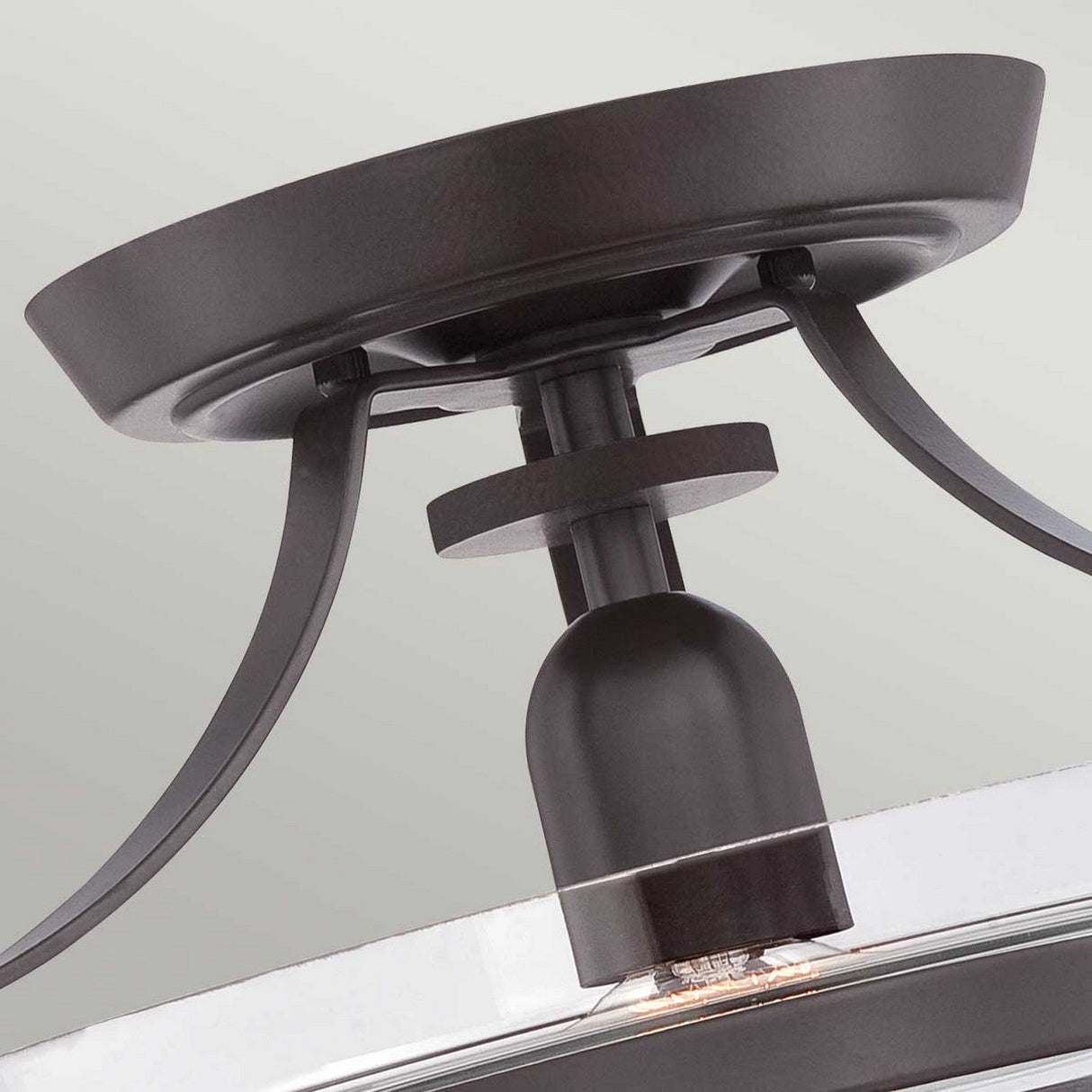 A close-up view of the Penn Station 1 Light Semi-Flush in Western Bronze showcases a ceiling light fixture featuring a dark metallic finish and an exposed bulb, highlighting its vintage industrial charm. Supported by gracefully arched metal arms connected to a circular base, this piece beautifully contrasts with the neutral gray background.