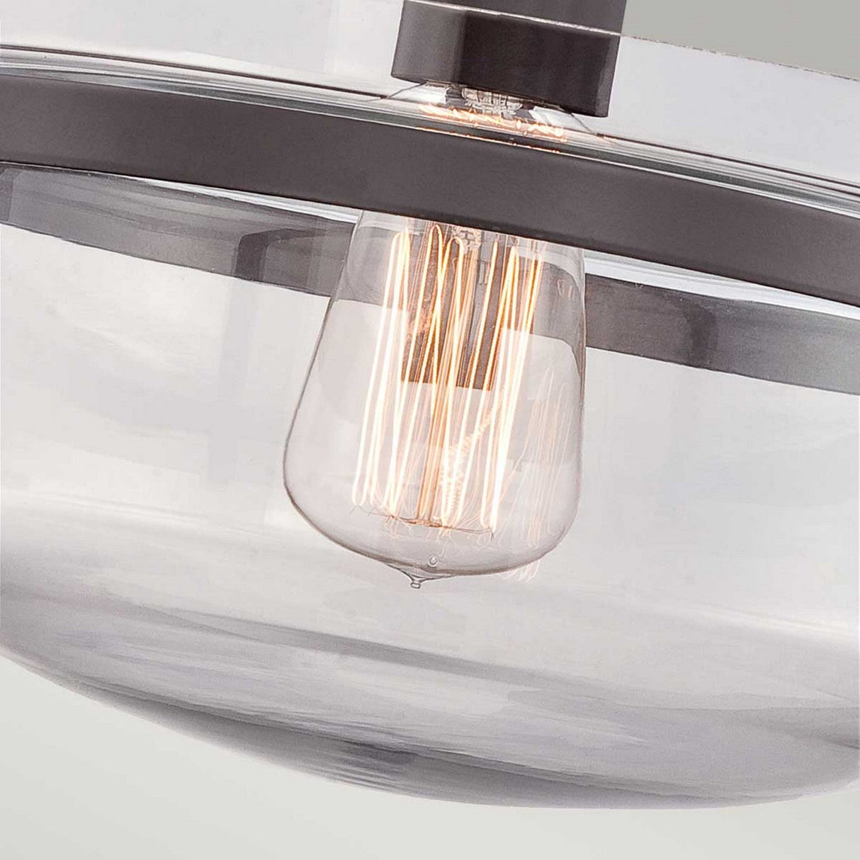 A close-up of the Penn Station 1 Light Semi-Flush in Western Bronze showcases its modern design with a clear glass shade and exposed filament bulb, emitting a warm glow. The sleek metal detailing adds a vintage industrial charm to the minimalist aesthetic.