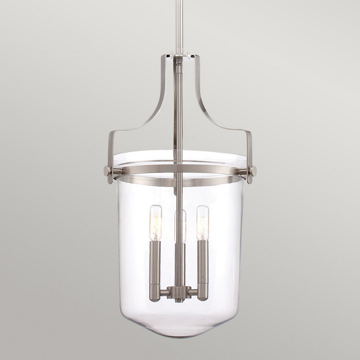 The Penn Station 3 Light Chandelier - Brushed Nickel exudes modern industrial elegance, boasting a brushed nickel frame and a clear cylindrical glass shade that encloses three vertical light bulbs, elegantly suspended against a neutral gray backdrop.