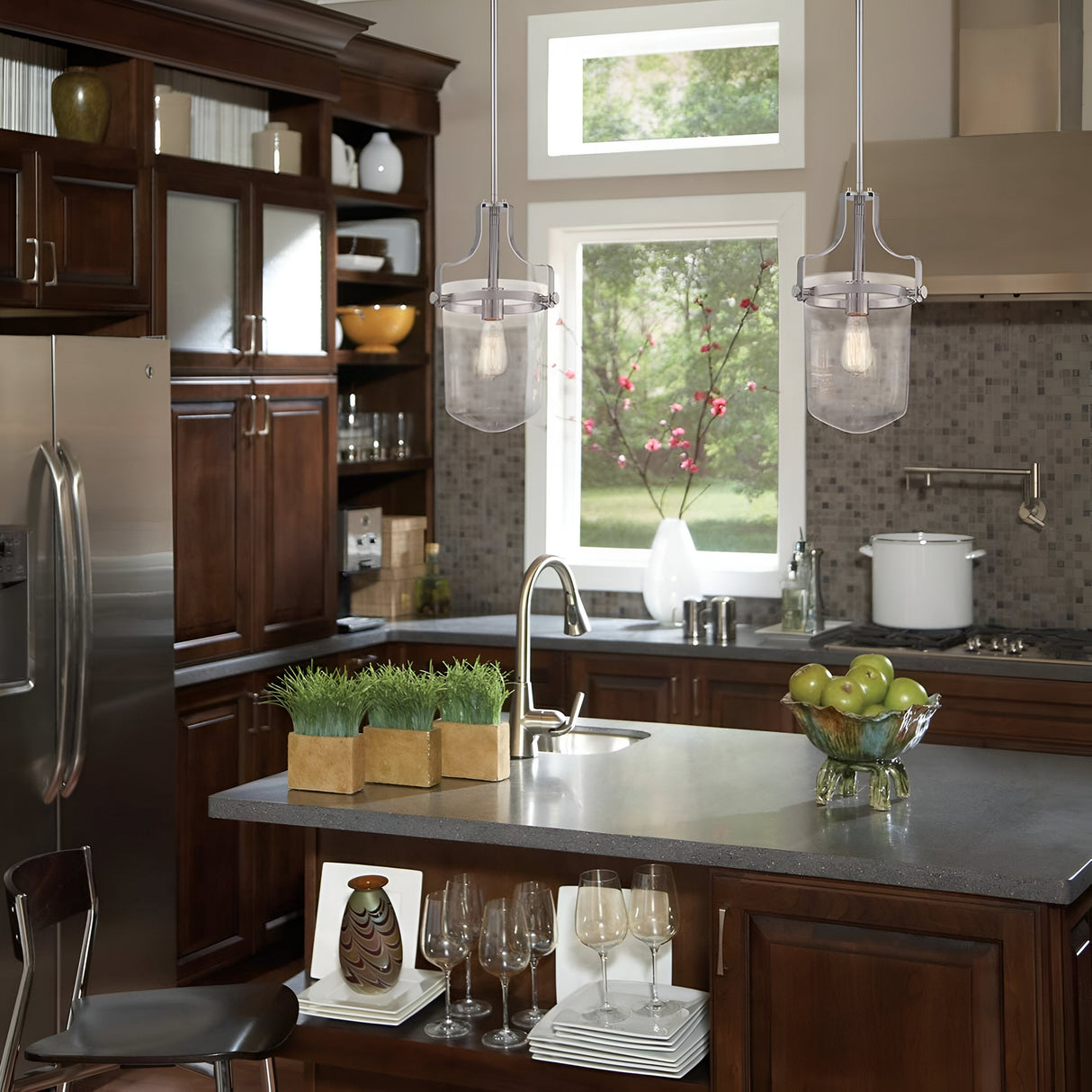 A modern kitchen with dark wood cabinets and stainless steel appliances exudes industrial elegance. A tile backsplash complements the brushed nickel accents. The Penn Station 3 Light Chandelier in brushed nickel illuminates two pendant lights hanging over a gray island countertop with a sink, where a bowl of green apples and a decorative grass plant sit stylishly.