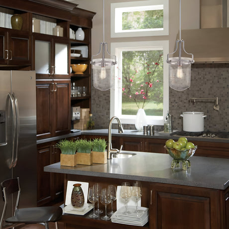 A modern kitchen with dark wood cabinets and stainless steel appliances exudes industrial elegance. A tile backsplash complements the brushed nickel accents. The Penn Station 3 Light Chandelier in brushed nickel illuminates two pendant lights hanging over a gray island countertop with a sink, where a bowl of green apples and a decorative grass plant sit stylishly.