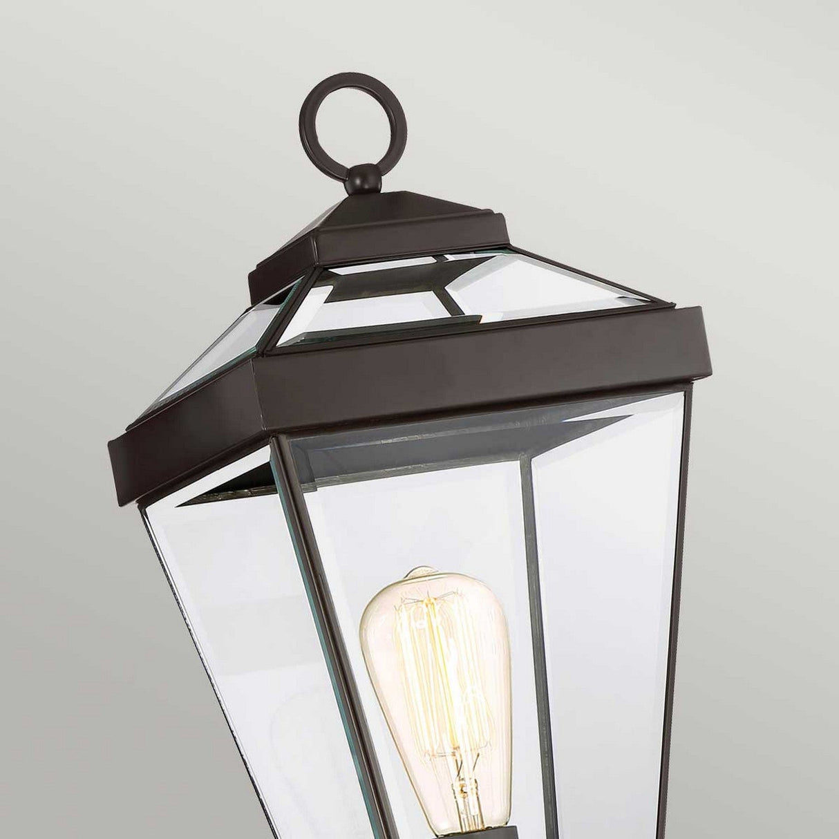 A close-up of the Ravine Medium Pedestal - Western Bronze features a black-framed, glass-paneled outdoor lantern with clear beveled glass showcasing a vintage-style bulb. The octagonal shape and Western Bronze finish add elegance, while the loop on top enhances its charm against a light gray background.