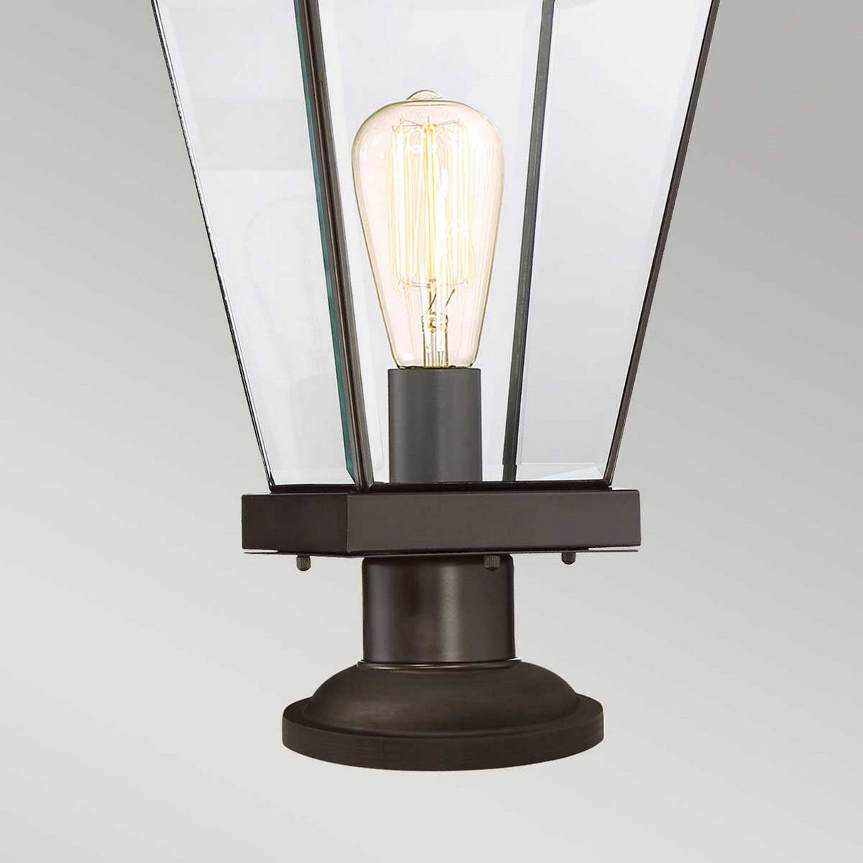 The Ravine Medium Pedestal - Western Bronze elegantly showcases a vintage-style light bulb within its lantern-like fixture, featuring angled clear beveled glass panels, as it sits against a light gray background.