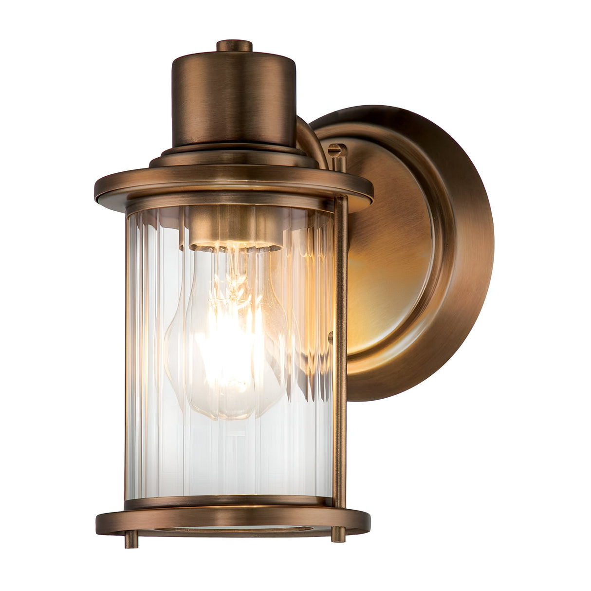 Introducing the Riggs 1 Light Wall Light in Weathered Brass, a wall-mounted sconce with a vintage appeal. It features a cylindrical glass cover encasing a glowing bulb and is crafted with an industrial style. The fixture includes a round backplate and metallic frame, making it an ideal choice for bathroom lighting.