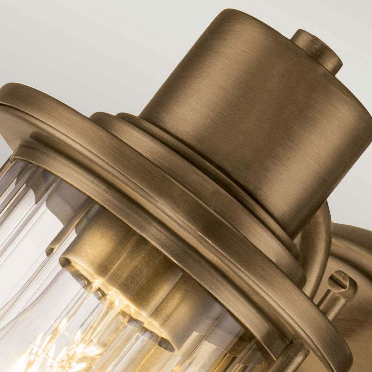 Close-up image of the Riggs 1 Light Wall Light in weathered brass, featuring a classic and elegant design with a clear ribbed glass shade. The visible bulb inside casts a warm glow across the metal surface, making it an ideal choice for adding an industrial style to bathroom lighting.