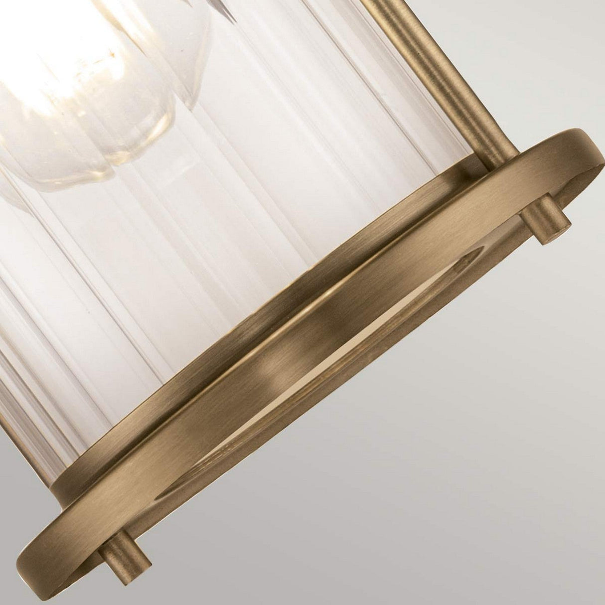 Close-up of the Riggs 1 Light Wall Light, showcasing a cylindrical glass shade with ribbed texture, encased in weathered brass-colored metal. Ideal for industrial-style bathroom lighting, the exposed bulb emits a soft glow through the transparent glass.