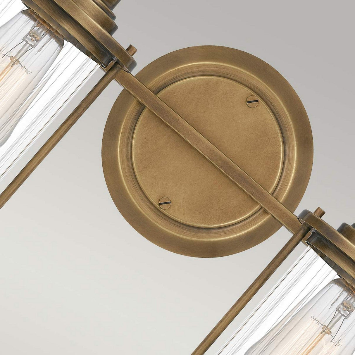 Close-up of the Riggs 2 Light Wall Light in weathered brass, featuring a round base and two ribbed glass cylinders. The wall light embodies industrial aesthetics with exposed light bulbs, set against a light gray background.