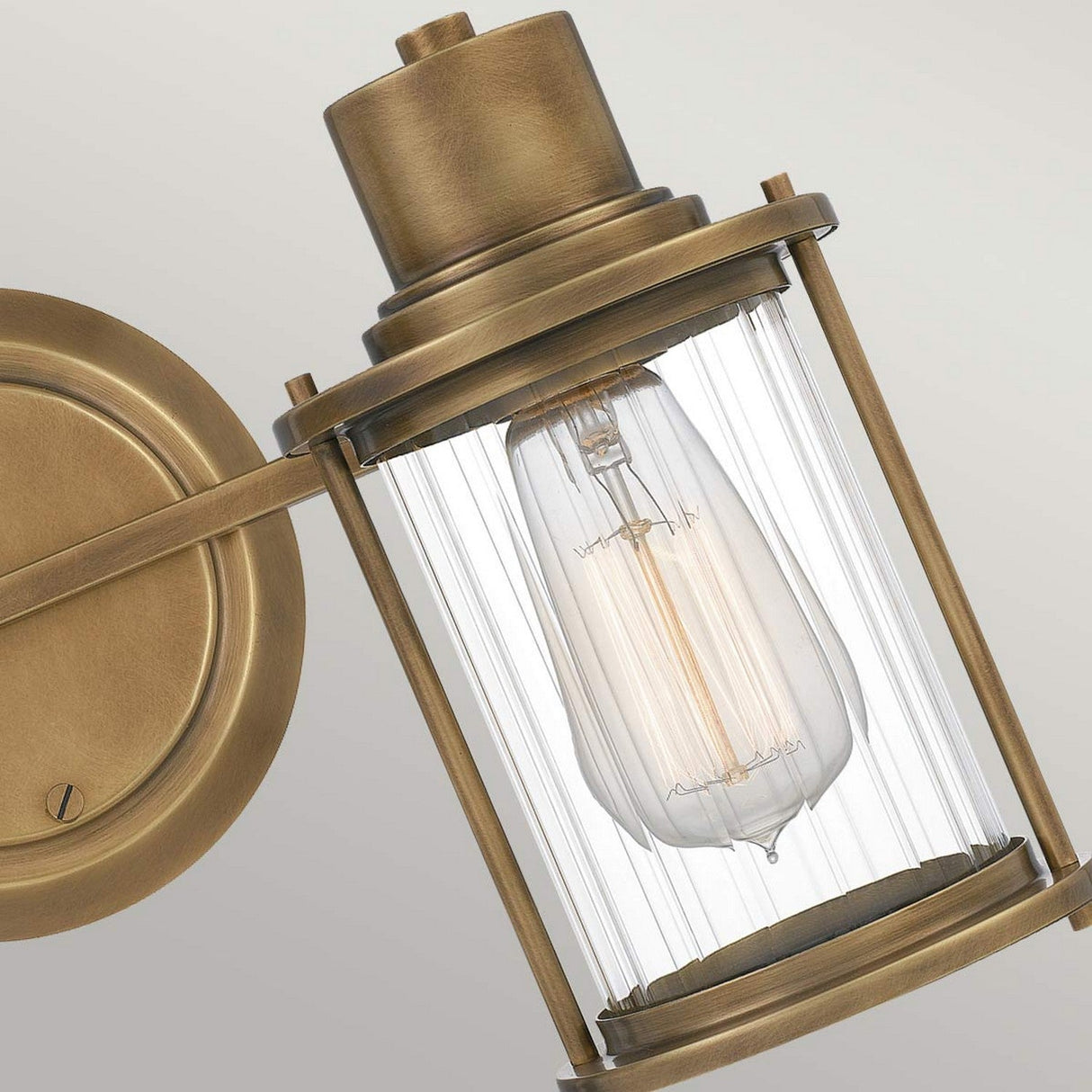 A close-up of the Riggs 2 Light Wall Light in a vintage style, featuring a weathered brass finish and a ribbed glass cylindrical shade. Inside, classic Edison bulbs are visible, casting a warm and inviting glow that embodies industrial aesthetics.