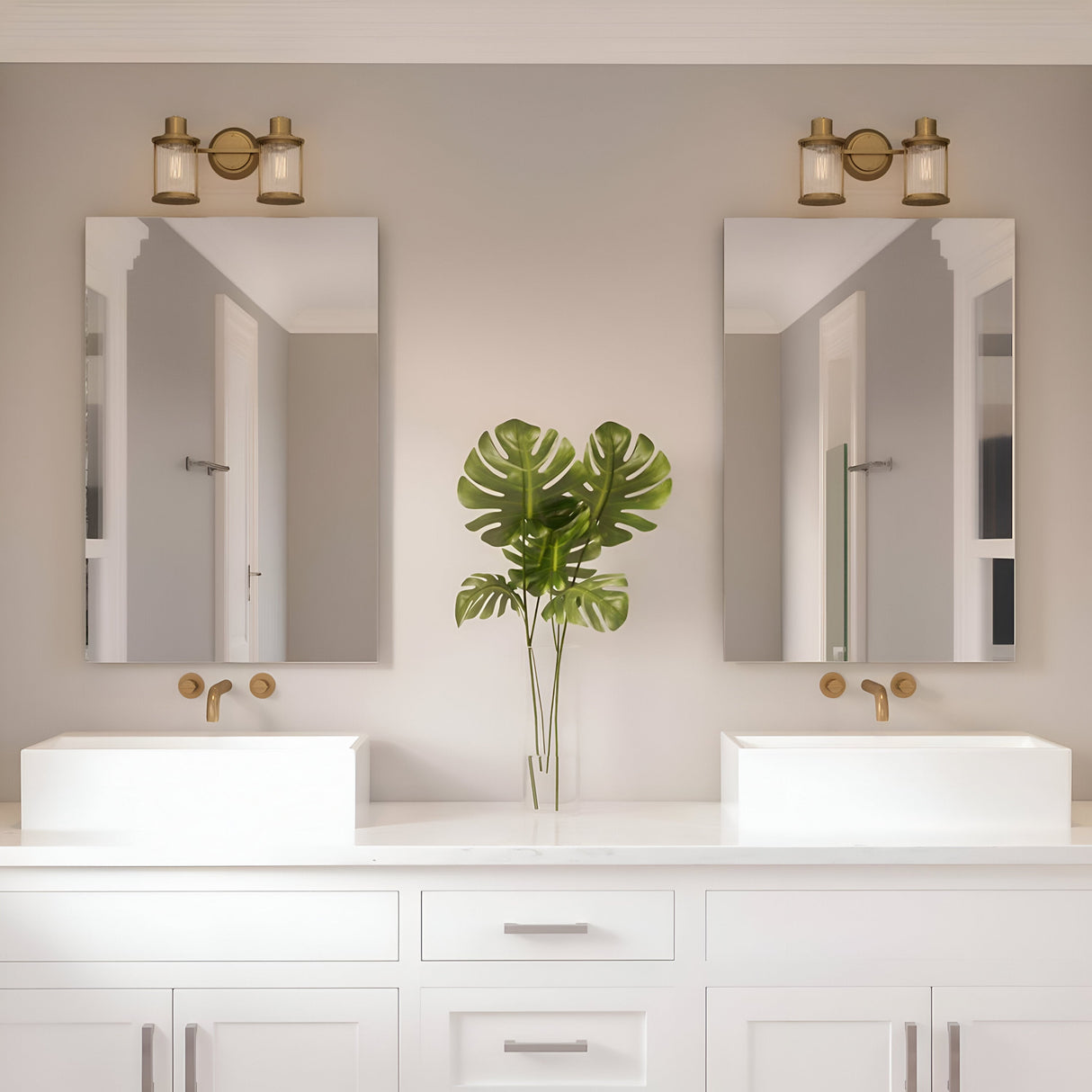 In a contemporary bathroom, two rectangular mirrors are positioned above white vessel sinks, each fitted with a weathered brass faucet. A large green monstera leaf stands between the sinks, while the Riggs 2 Light Wall Light in Weathered Brass provides a touch of industrial flair with its ribbed glass fixtures mounted overhead.