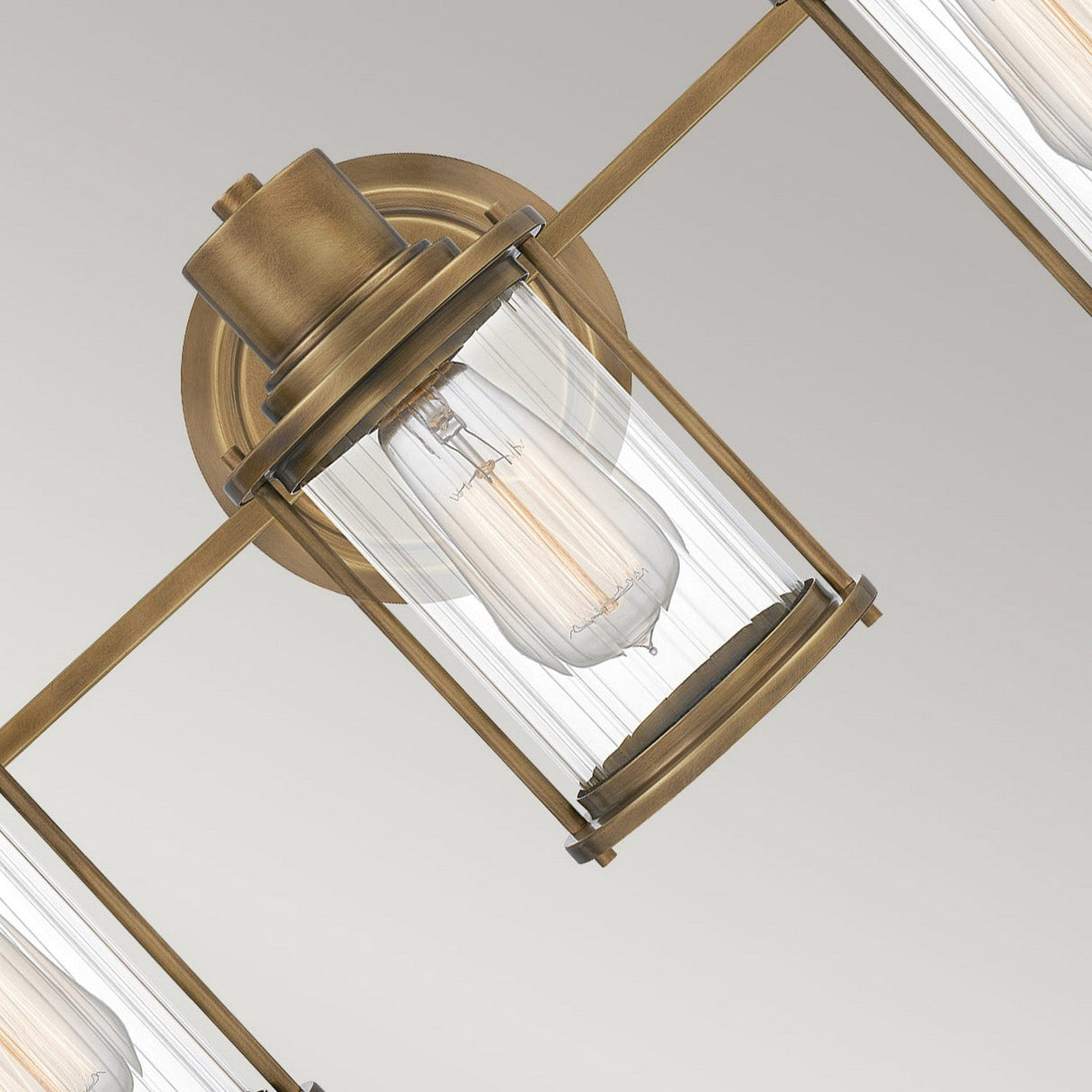 Close-up of the Riggs 3 Light Wall Light in weathered brass, showcasing its vintage-style design with an exposed bulb within a cylindrical, ribbed glass and metal fixture. The light is secured on a matching round base, adding sleek elegance perfect for bathroom lighting.
