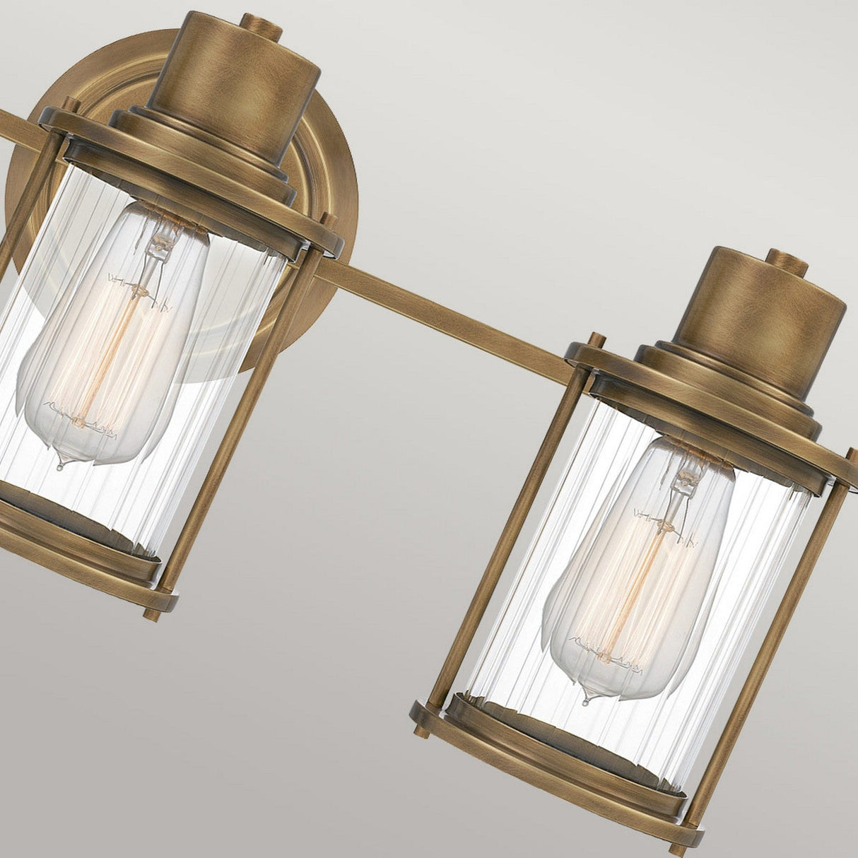 A detailed view of the Riggs 3 Light Wall Light in weathered brass, featuring ribbed glass shades that display exposed Edison-style bulbs, set against a light grey background.