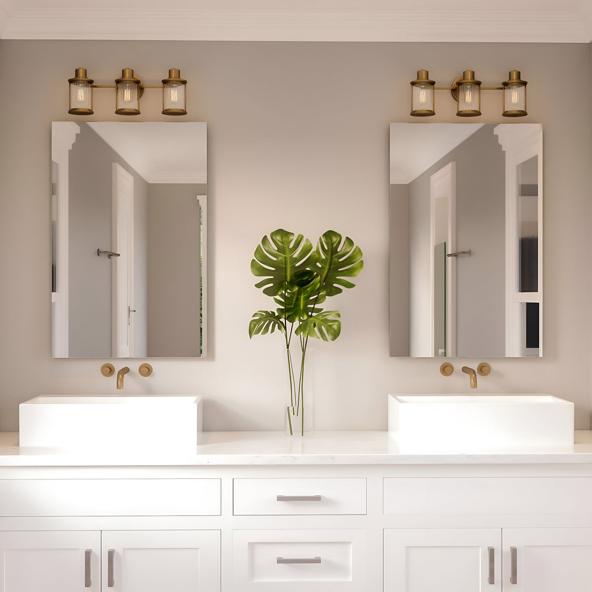 A modern bathroom showcases dual white sinks on a countertop, each accompanied by a rectangular mirror. The Riggs 3 Light Wall Light in weathered brass, with its ribbed glass shades, highlights the faucets and light fixtures, perfectly complementing the neutral decor. A tall vase with a large leafy plant is centered between the sinks.