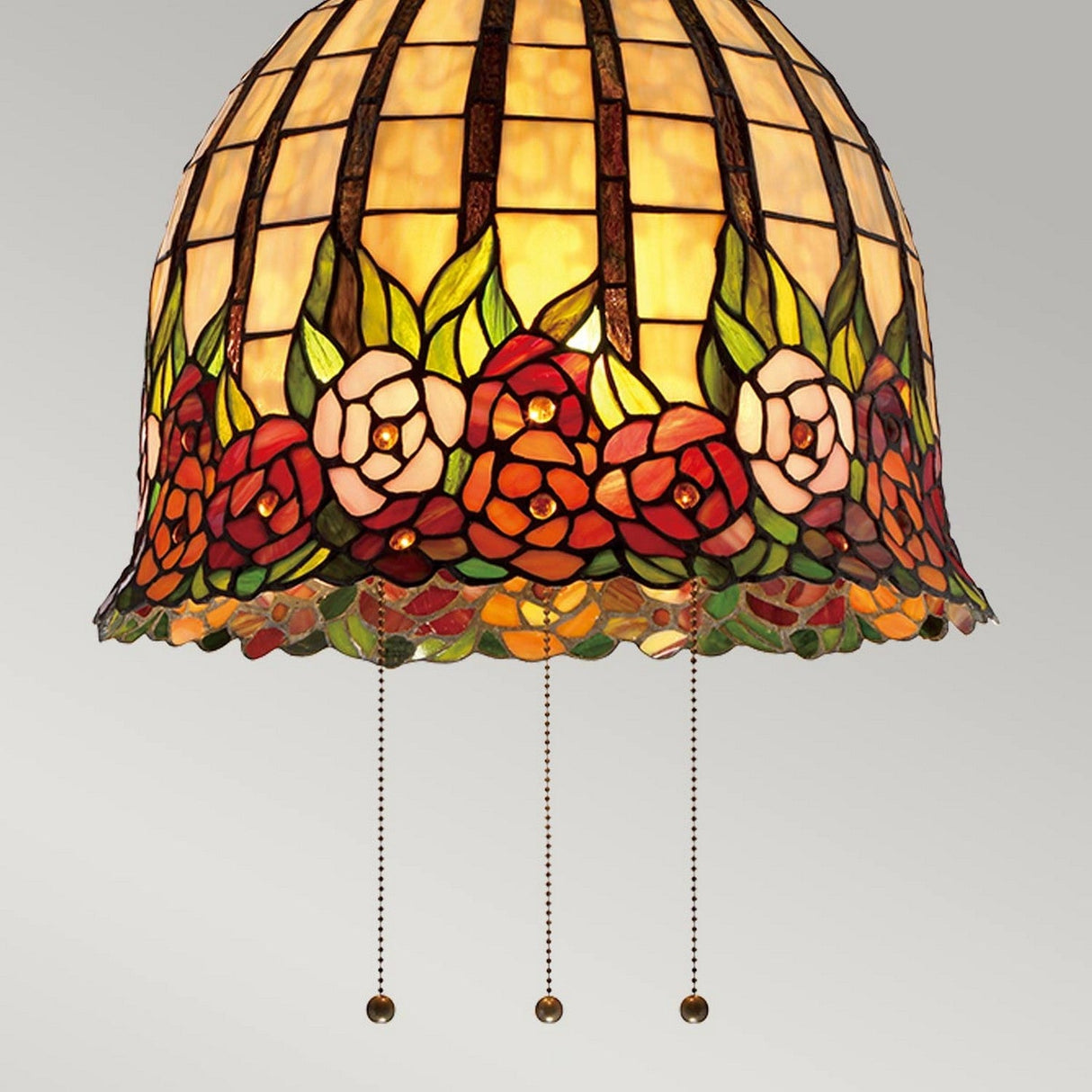 The Rosecliffe 3 Light Tiffany Pendant in an Imperial Bronze finish showcases a dome-shaped stained glass lampshade, beautifully adorned with a floral design of red, pink, and white roses complemented by green leaves. Vivid light shines through the colorful rose-patterned glass panels, enhanced by three pull chains hanging below.