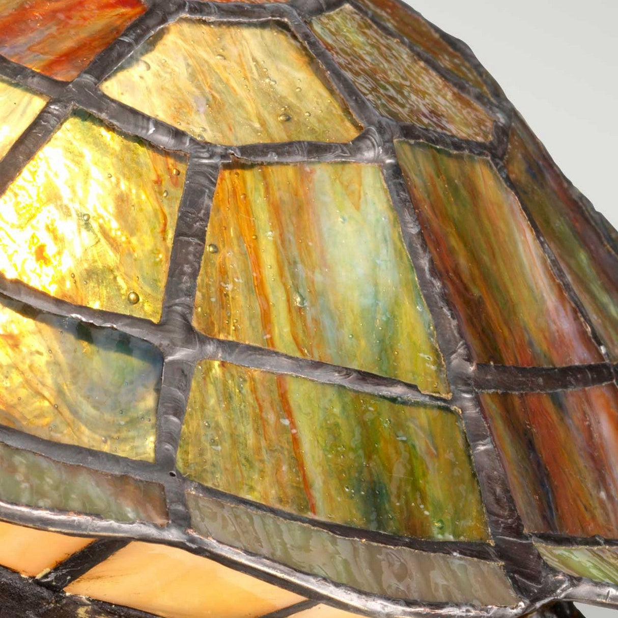 Close-up of the Tiffany Animal Lamps Sawback Turtle Lamp - Vintage Bronze, showcasing a stained glass turtle shell pattern. The glass displays amber, green, and orange hues, framed by dark lead around each piece to form an intricate mosaic design.