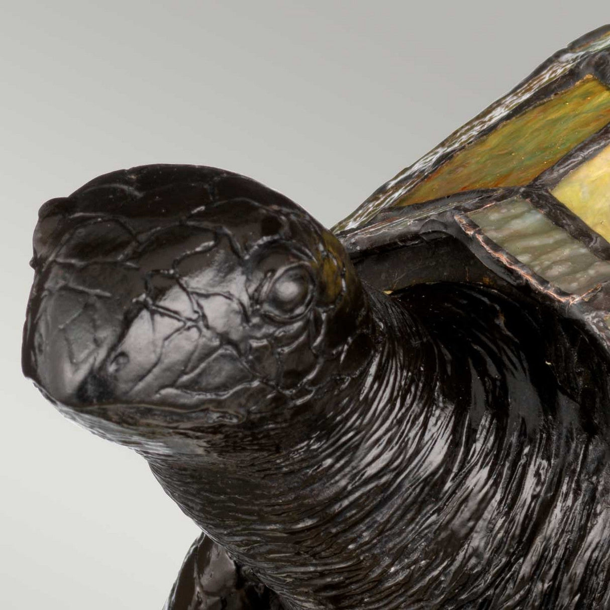 A close-up of the Tiffany Animal Lamps Sawback Turtle Lamp - Vintage Bronze highlights its intricate detailing on the textured shell and head, reminiscent of the artistry seen in stained glass pieces, set against a plain, light gray background.
