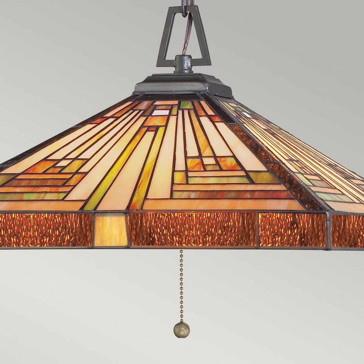 The Stephen 3 Light Tiffany Pendant in Vintage Bronze showcases Arts & Crafts influence with geometric stained glass patterns in shades of amber, orange, and cream. It features a pull chain for operation and is elegantly set against a neutral gray background.