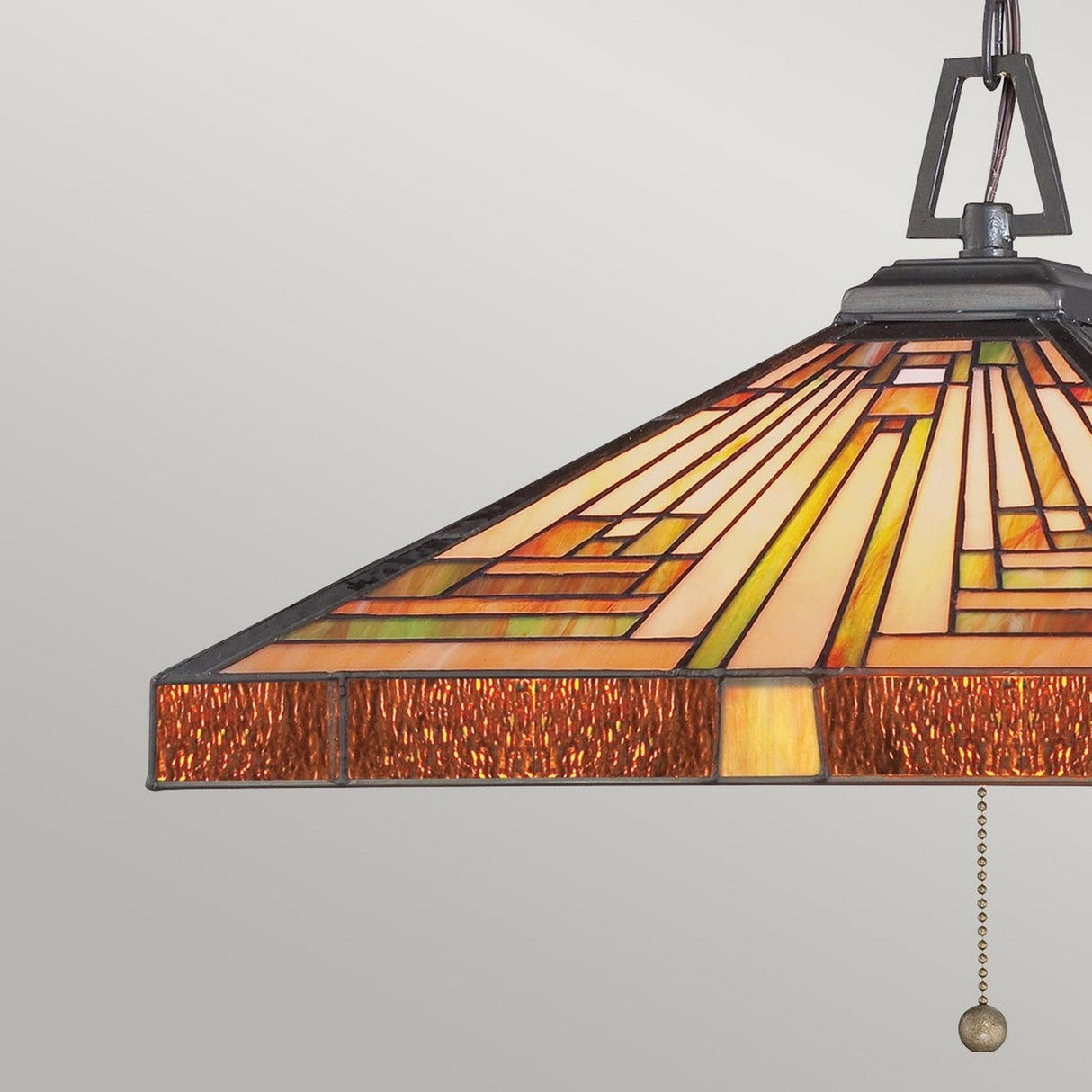 The Stephen 3 Light Tiffany Pendant in Vintage Bronze showcases a geometric stained glass design with shades of amber, orange, and yellow and is influenced by the Arts & Crafts style. It features a black metal frame and a pull chain set against a plain gray background, exuding vintage elegance.