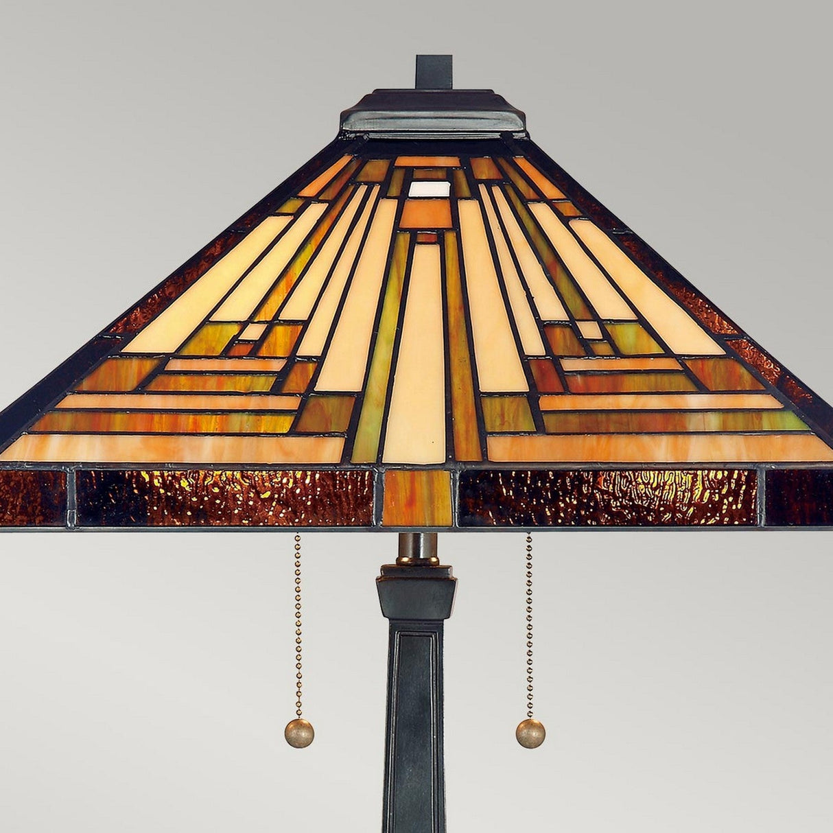 Presenting the Stephen 2 Light Tiffany Table Lamp in Vintage Bronze, featuring a stained glass shade with geometric patterns in warm shades of amber, brown, and cream. The lamp's metal base is elegantly crafted and includes two pull chains with small round weights.