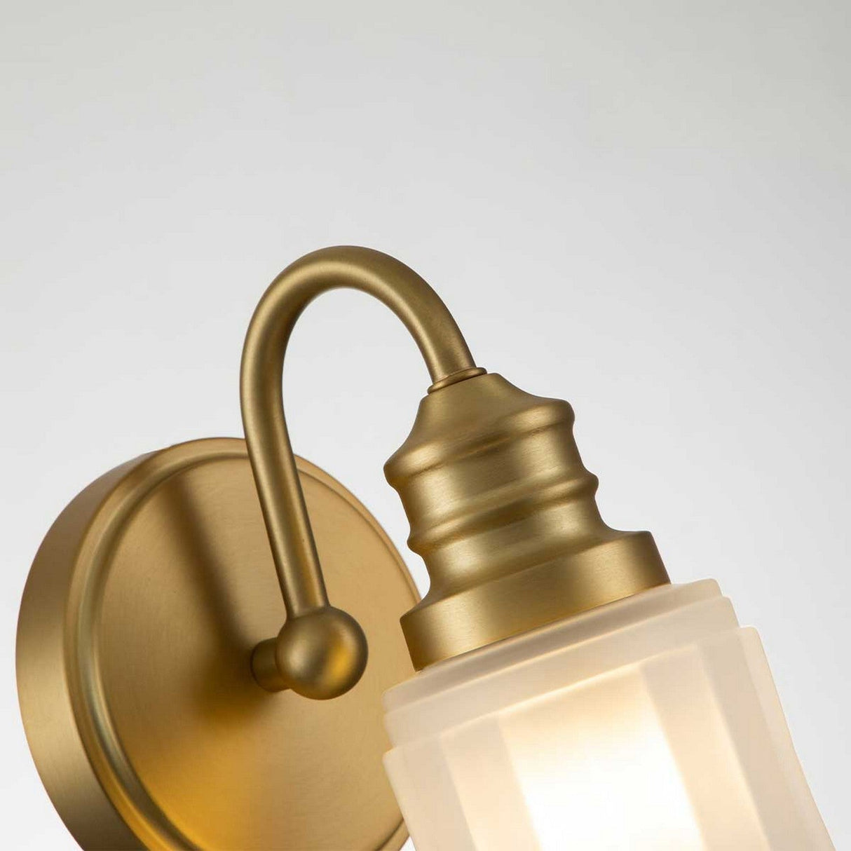 The Swell 1 Light Wall Light in Brushed Brass features a frosted glass shade that provides ambient illumination, complemented by an elegant curved arm and round base for a classic and sophisticated design.