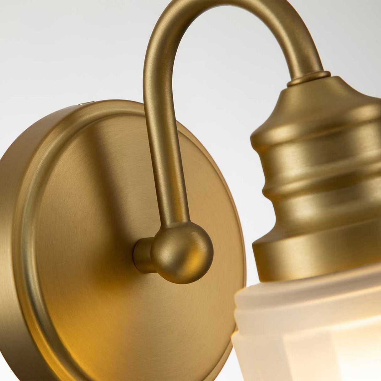 Close-up of the Swell 1 Light Wall Light - Brushed Brass, featuring a wall-mounted design with a frosted glass shade. This light fixture highlights a curved arm and smooth, brushed finish, offering ambient illumination to any space.