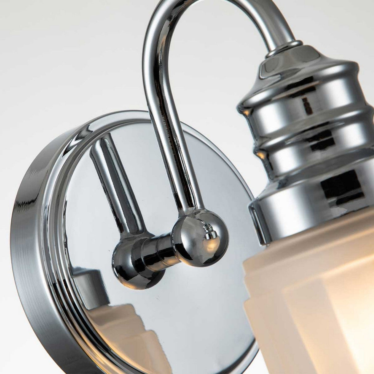 Close-up of the Swell 1 Light Wall Light - Polished Chrome, featuring a curved arm and round base finished in silver chrome. Boasting an IP44 rating, this fixture has a frosted glass shade that provides an elegant and contemporary appearance against a light background.