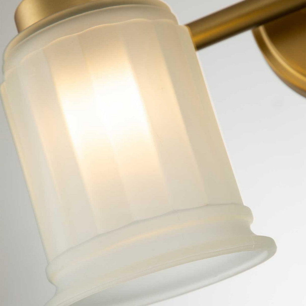 Close-up of the Swell 2 Light Wall Light in brushed brass, showcasing its arm and frosted glass shade. The fixture gently diffuses light through the textured ripples of its cylindrical glass against a plain white wall.