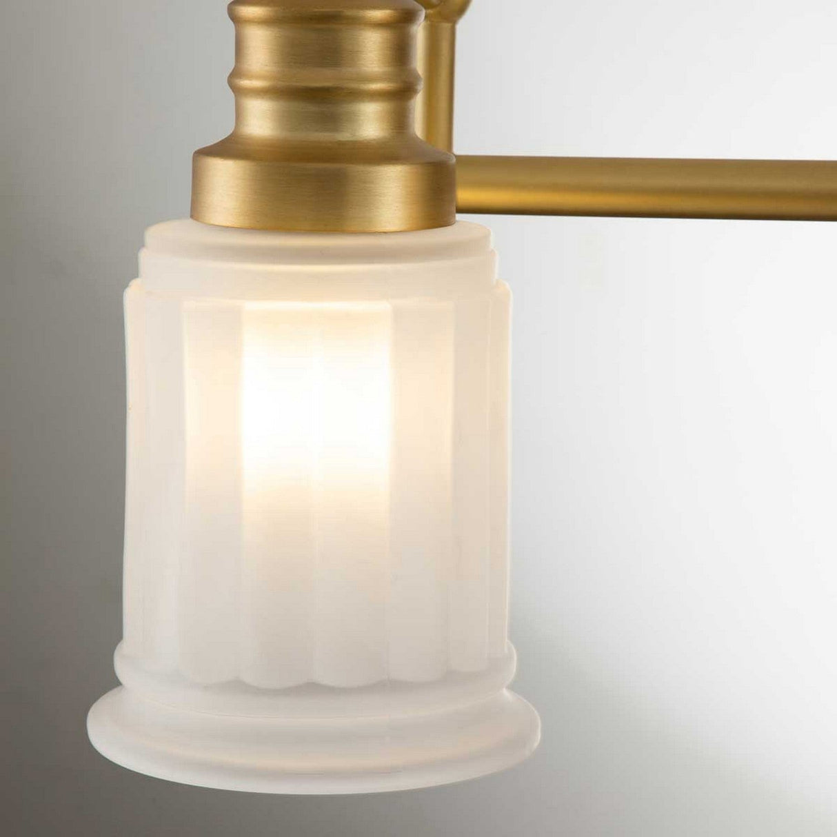 A close-up of the Swell 3 Light Wall Light - Brushed Brass showcases an elegant light fixture with a frosted glass shade featuring vertical grooves, complemented by its sophisticated brushed brass finish. The LED G9 bulb softly glows through the glass, adding to its charm.