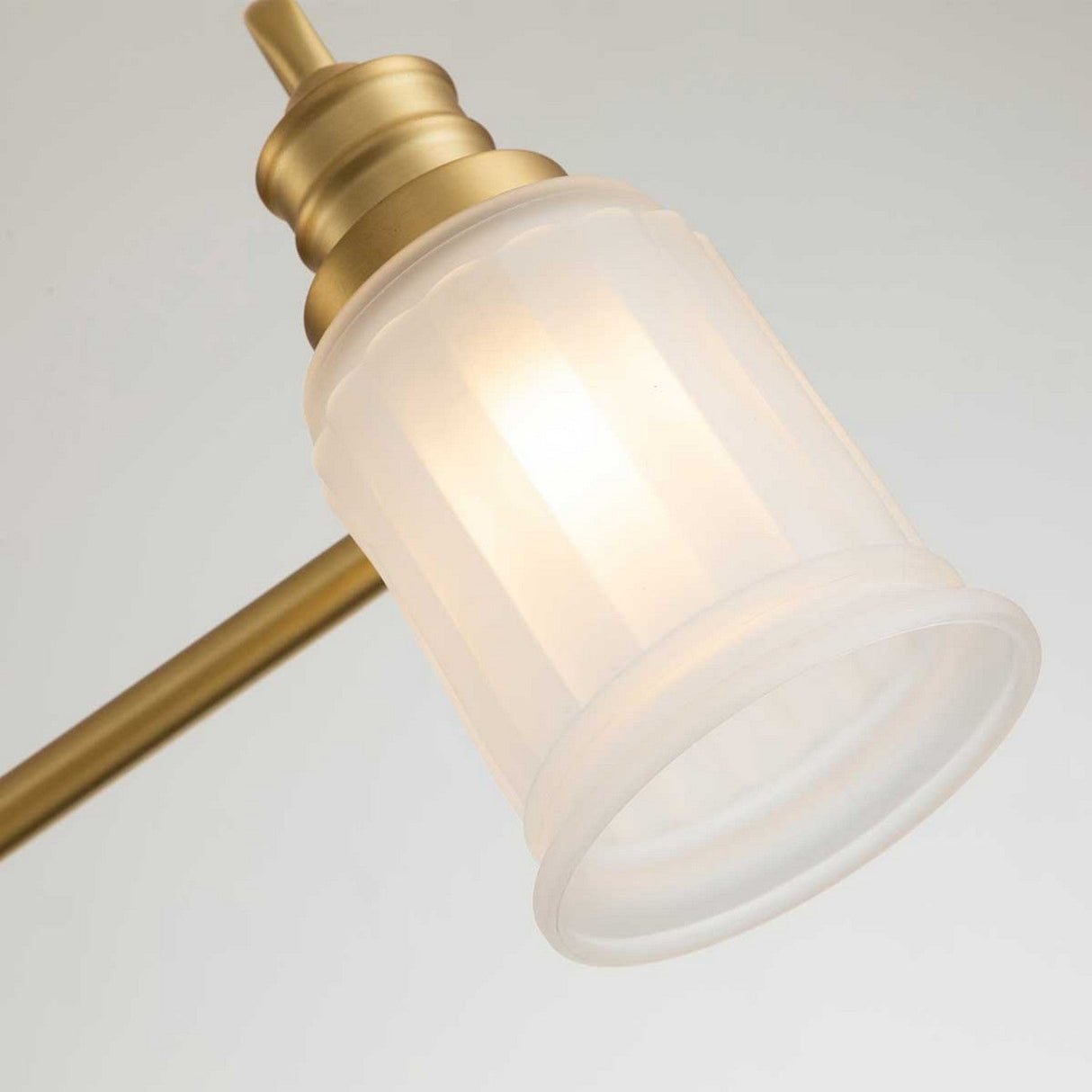 A close-up of the Swell 3 Light Wall Light in brushed brass showcases its frosted glass shade. The LED G9 light is illuminated, casting a warm glow. With its classic cylindrical shape and metal accents, this fixture adds an elegant touch against a neutral, light-colored backdrop.