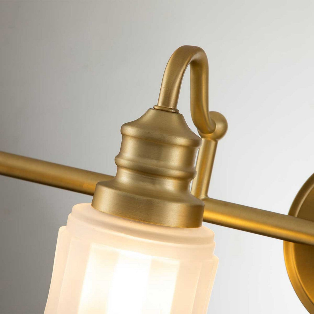 Close-up of the Swell 4 Light Wall Light in brushed brass, featuring a frosted glass shade. The light is illuminated, casting a warm glow that showcases metropolitan sophistication. With its curved arm and elegant detailing, this fixture is perfect for bathroom vanity lighting.