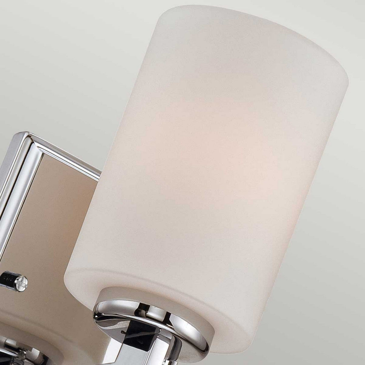 A detailed shot of the Taylor 1 Light Small Wall Light in polished chrome highlights its sleek design, featuring an opal glass cylindrical shade and a shiny chrome base. The light gray background amplifies the fixture's contemporary allure.