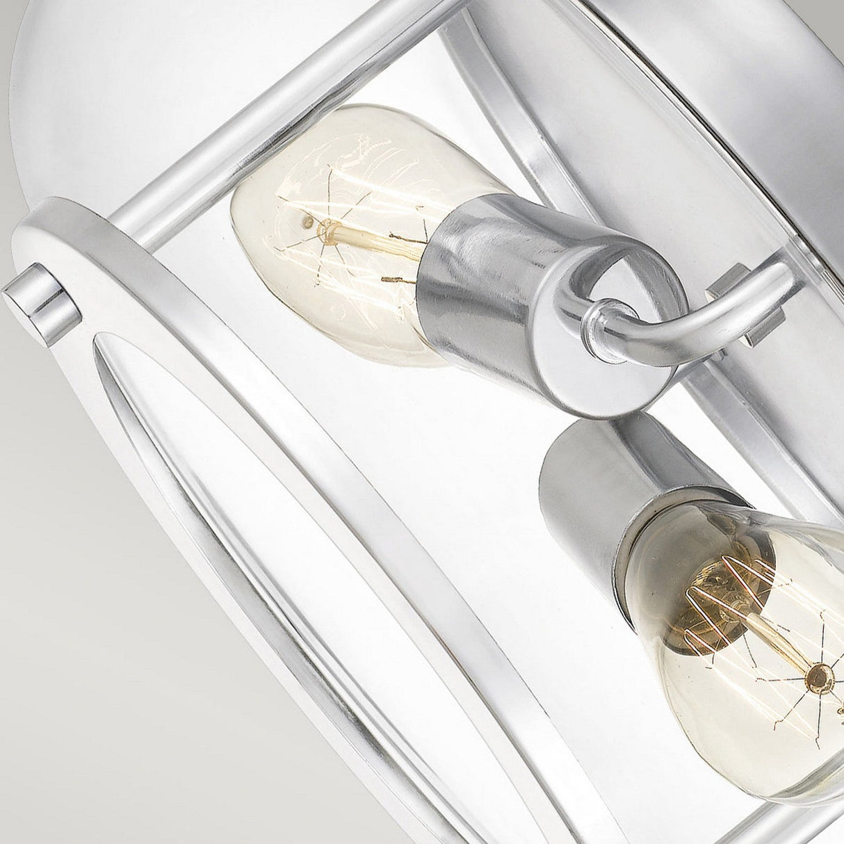 A close-up of the Trilogy 2 Light Flush in polished chrome reveals two vintage-style light bulbs with visible filaments nestled within a metallic fixture. The sleek, circular design highlights modern industrial style, accentuated by the polished chrome details against a neutral background.