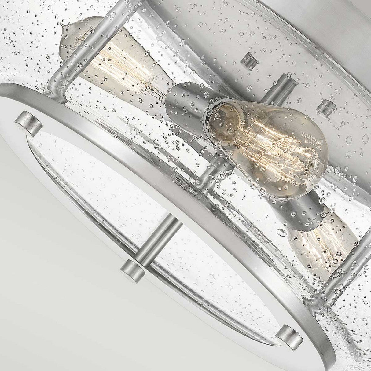 The Trilogy 3 Light Flush - Brushed Nickel ceiling light fixture features a modern design with a circular seeded glass shade adorned with small bubbled textures. Inside, two visible Edison-style LED bulbs are enhanced by a sleek metal frame and hardware in brushed nickel.