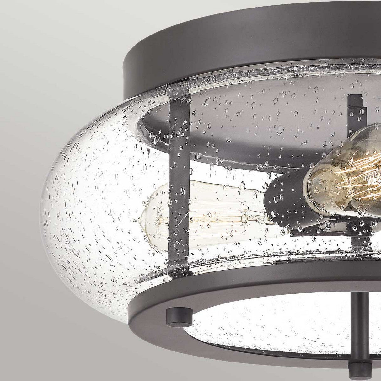 A close-up of the Trilogy 3 Light Flush - Old Bronze reveals a modern ceiling light fixture with a seeded glass shade adorned with small bubbles. Inside, two Edison-style LED filament bulbs are encased within an Old Bronze frame, showcasing a fusion of contemporary and industrial design elements.