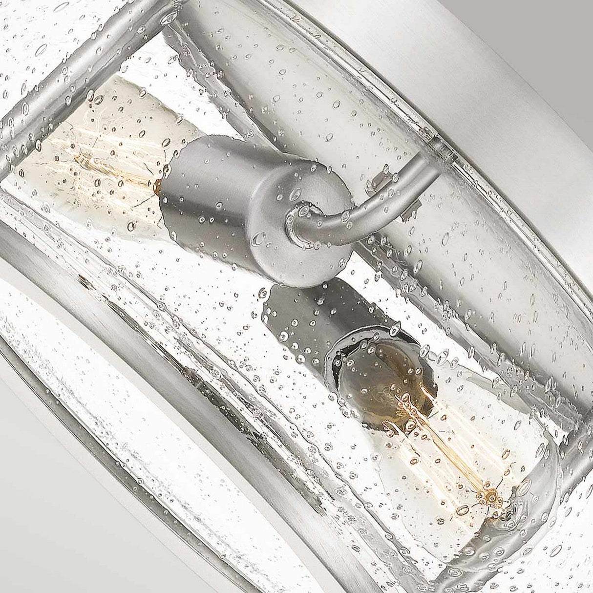 Close-up of the Trilogy 2 Light Flush - Brushed Nickel, showcasing two exposed bulbs within textured, seeded glass. Its sleek finish adds a modern touch to the design, enhancing the contemporary aesthetic with stylish flair.